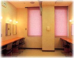 壁の両側に机や椅子、鏡が設置され、うす紫色のブラインドが窓から降ろされている洋室の楽屋の写真