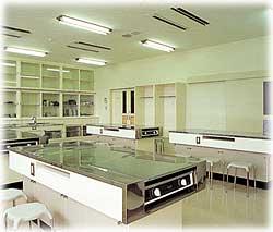 ガス台が複数調理台に設置され、壁にはガラス棚が据え付けられている白やベージュを基調とした清潔感のある調理実習室の写真