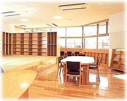 明るい色のフローリングに白いテーブルとダークオーク色の椅子が置かれ、奥には一段高くなった床で座って本を読めるような場所、周りには本棚が壁一面に敷き詰められていて窓から明るい日差しが差し込む図書室の写真