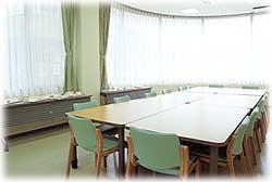 中央に複数のテーブルが一つにまとめられ、周りに若草色の椅子が据え付けられている、大きい窓から日差しが差し込む明るい研修室（洋室）の写真
