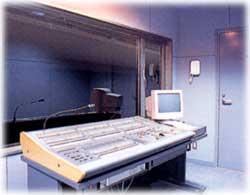大きな音響設備や収音スペースがある薄い青紫色の照明で照らされた音響調整室の写真