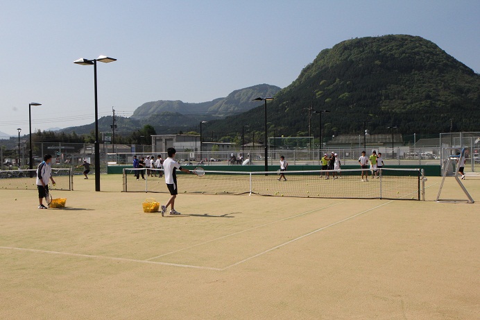 運動着を着た人達が、大きな山の麓にあるテニスコートで練習している写真