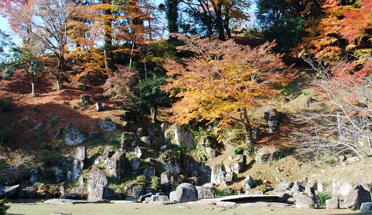 紅葉に彩られた庭園の山に、形の違う石で形作られた枯瀧が組まれている藩主御殿庭園の写真