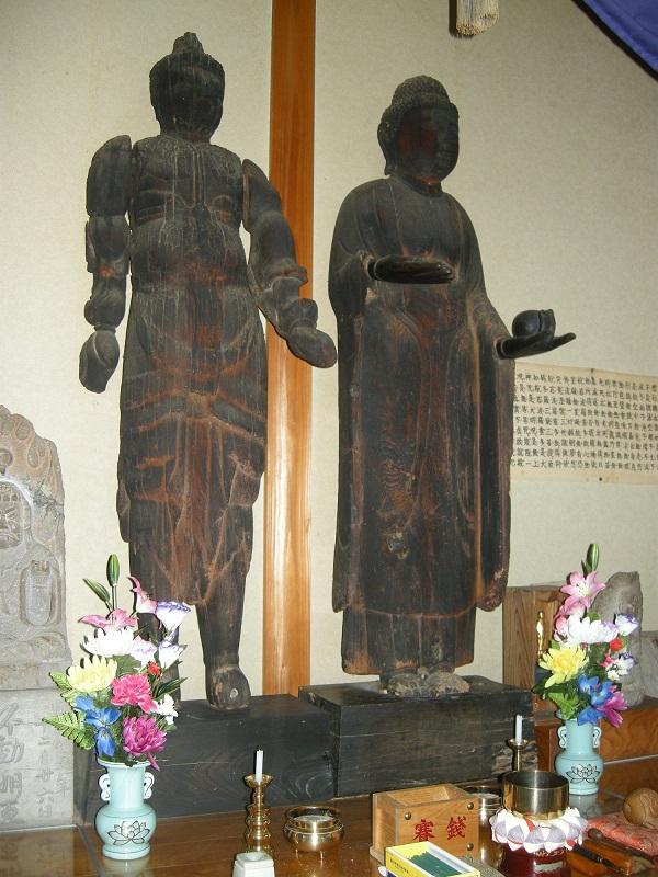 左側に左手が少し斜め上に上がり直立の毘沙門天像、右側は薬壷を左手に持ち、右手の手のひらを胸の高さまで上げている薬師如来像の2体の像が横長の木材の土台の上に設置されている写真