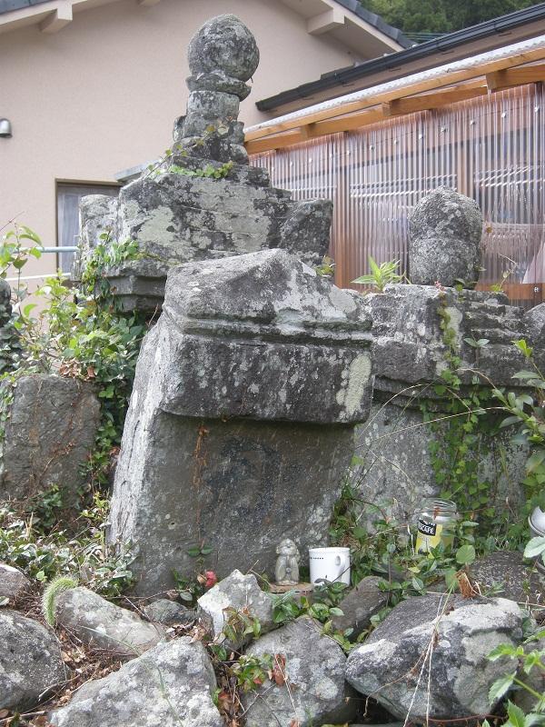 周囲に崩れ落ちた石が散乱したり、形の違う2つの石塔の前に、頂上が山型で造られ二重の切込みが施されている板碑が斜めに立っている写真