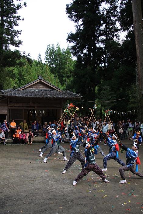 神社の拝殿の前の境内で、額に白色の鉢巻のようなものを巻き、上は青色の着物の形、下はもんぺを履いた男の子たちが2名ずつ向き合って、長い棒を交差させて舞っている大浦楽の写真