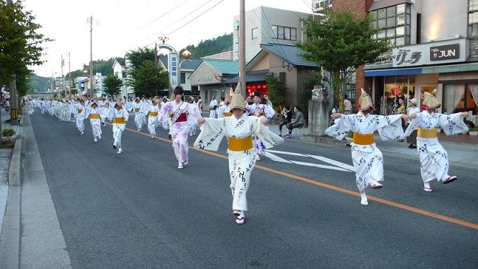 道路に横3列になり、山吹色の帯を締め白色の浴衣を着た女性たちが山路踊りを踊っている様子の写真