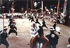 亀都起神社の境内で、全身黒色の衣装に身を包み、頭に白色の鉢巻のようなものを巻いて、棒杖を両手で持って演じている山田楽の様子の写真