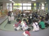 図書館内の広場で、エプロンを付けた女性が前で立って話しているのを、親子で参加している女性と子どもたちが座って話しを聞いている写真