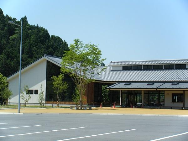 青空の下、中央に近代的な瓦屋根風道の駅の建物があり、左側に濃い緑の木々が生えた山、建物周辺には若葉色の木々が植えられ、全面には広々とした駐車場がある写真