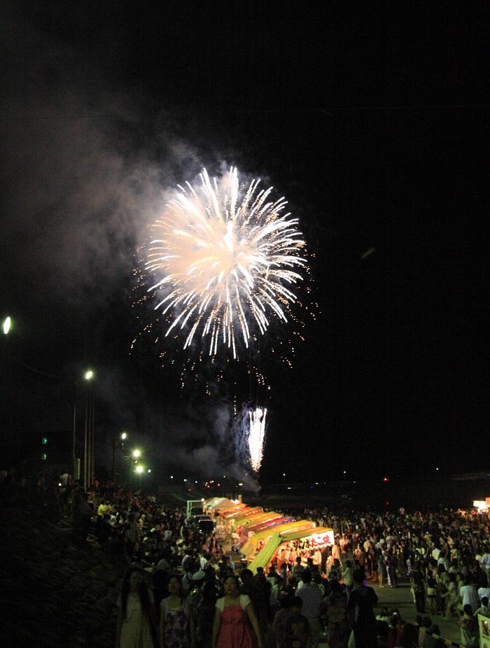 賑わう屋台と夏祭りに集まる人々の上空で花火が咲いている写真