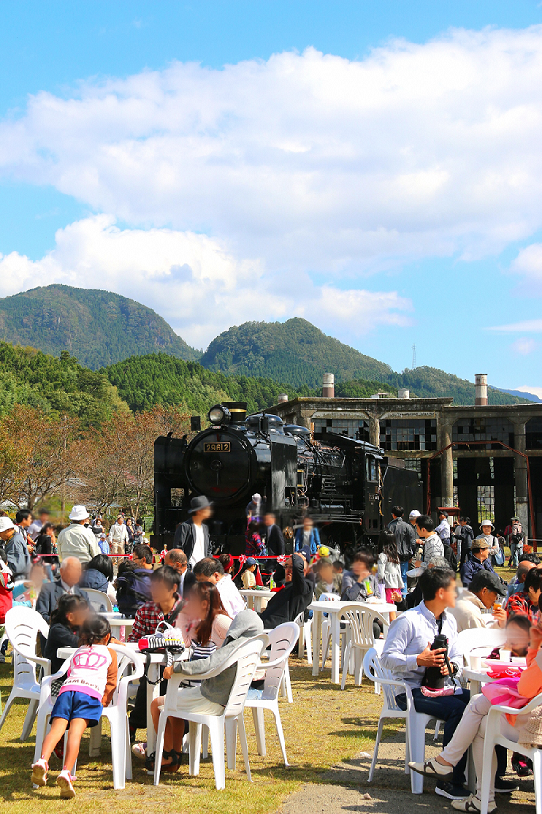 雲が流れる青空の下で緑の山々を背景に豊後森機関庫公園で蒸気機関車を眺める人たちやテーブルを囲み談笑する人々を撮影した写真